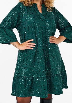 Femme portant une robe à sequins vert émeraude, manches trois-quarts, ourlet froncé, debout les mains sur les hanches devant un fond blanc.