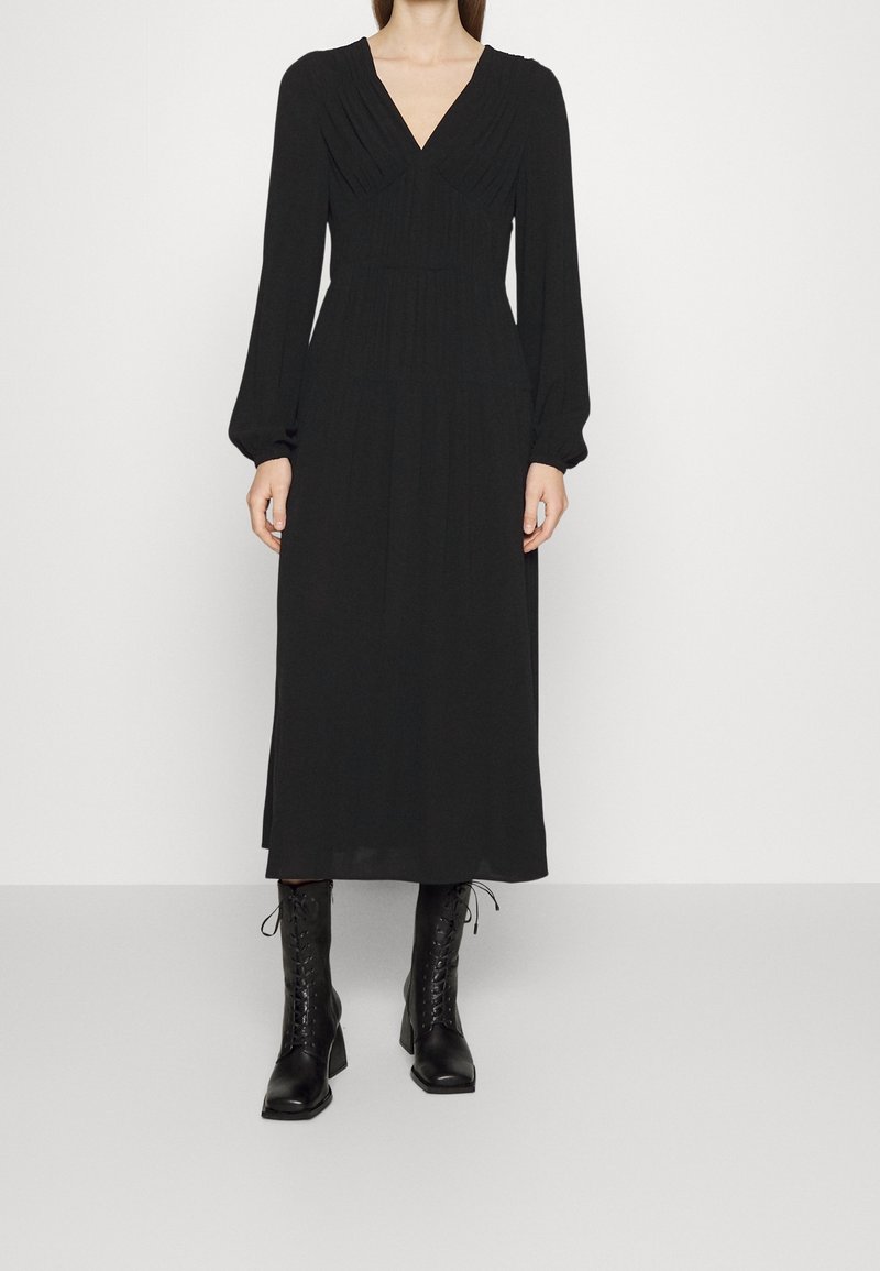 Femme portant une longue robe noire à manches longues et col en V, associée à des bottines noires à lacets à talons.