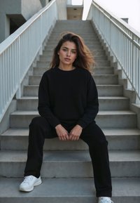 Sudadera negra y pantalones jogger, hechos de tela suave, combinados con zapatillas blancas. El modelo se sienta con confianza en escalones de concreto gris.