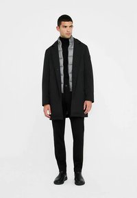 Abrigo negro sobre un chaleco acolchado gris, suéter de cuello alto negro y pantalones negros. Tejido liso con un ajuste entallado y diseño minimalista. Zapatos negros.