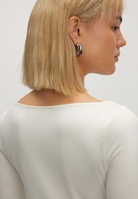 Witte longsleeve top met een brede ronde hals, gemaakt van gladde stof. Korte, rechte blonde haren met zilveren creolen.