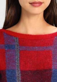 Frau, die einen roten Pullover mit blauem und grauem Karomuster trägt, von den Schultern bis zu den Lippen zu sehen, mit langen dunklen Haaren.