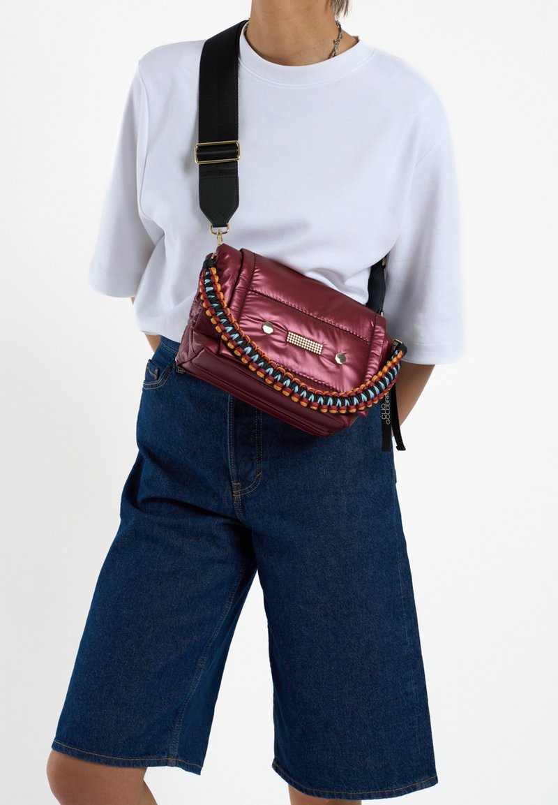 Bordeaux gekleed schoudertasje met een kleurrijke gevlochten schouderband, metalen accenten en ritssluiting. Gedragen over een wit shirt en denim shorts.