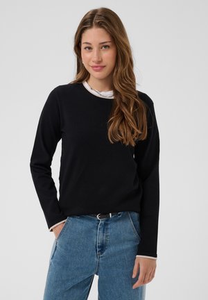 Jeune femme aux longs cheveux ondulés portant une chemise noire à manches longues sur un haut blanc, un jean bleu, les mains dans les poches, sur un fond uni.