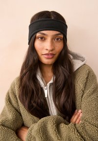 Crna runo traka za glavu nošena preko duge, tamne kose; uparen s sivom majicom s kapuljačom i maslinasto zelenom teksturiranom jaknom. Mekana tkanina, jednostavan dizajn.