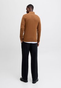 Maglione marrone a maglia con collo, indossato sopra una camicia a righe bianche, abbinato a pantaloni neri larghi e scarpe nere.