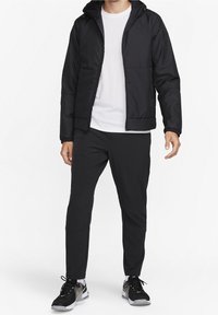 Homme portant une veste à capuche noire, un pantalon noir, un t-shirt blanc et des chaussures de sport noires et grises, debout sur un fond blanc.