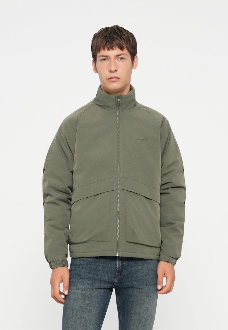 Chaqueta verde oliva con cuello alto, cierre frontal y dos grandes bolsillos delanteros. Presenta una textura suave y mate con costuras en contraste.