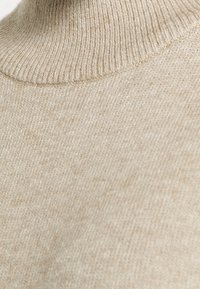 Beige höghalsettröja i mjukt stickat material. Har en ribbad struktur runt kragen och kroppen, med en slät yta.