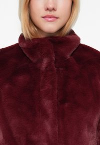 Abrigo de piel burdeos con textura suave, cuello alto y diseño sin costuras. Sin botones visibles ni detalles metálicos.