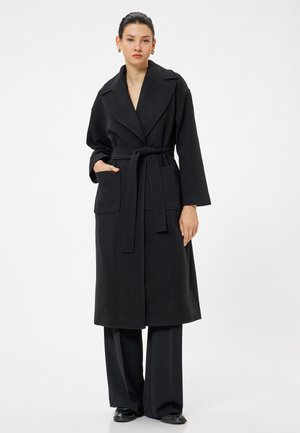Gabardina negra con cuello de chal, cinturón y bolsillos frontales. Hecha de una tela texturizada, crea una silueta larga y fluida.