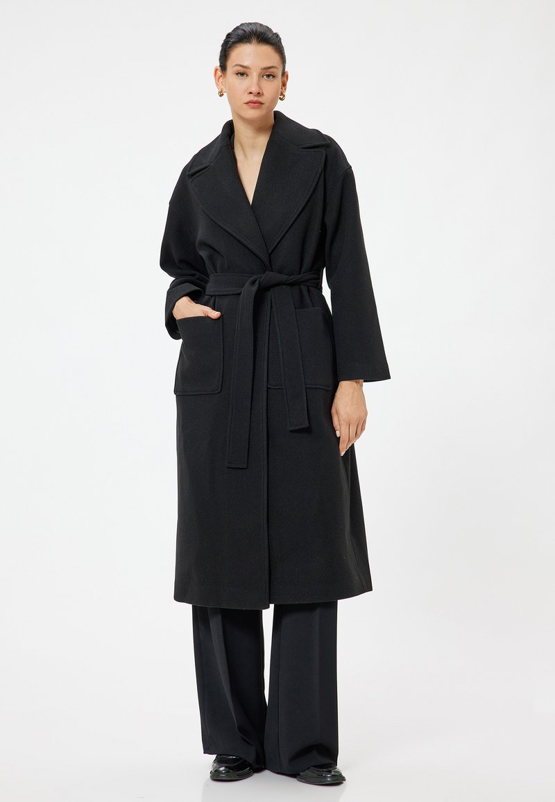 Cappotto nero con colletto a scialle, cintura e tasche frontali. Realizzato in tessuto testurizzato, crea una silhouette lunga e fluida.