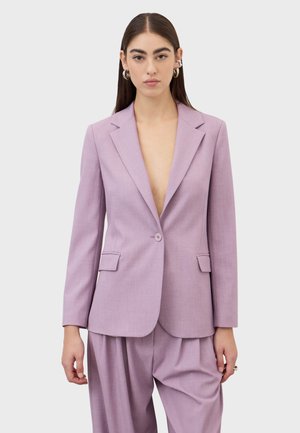 Donna con capelli lunghi e scuri che indossa un blazer su misura color viola chiaro e pantaloni a gamba larga abbinati, su sfondo grigio chiaro.