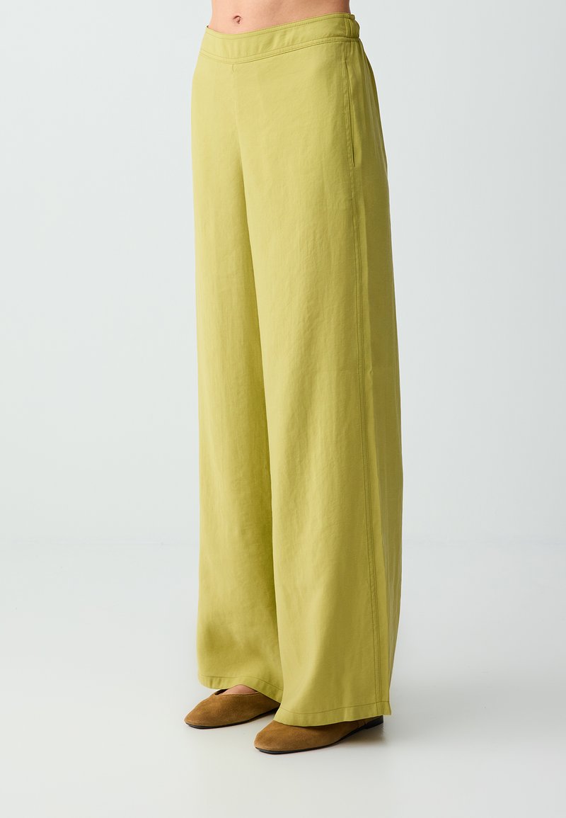 Femme portant un pantalon vert clair taille haute à jambes larges, associé à des chaussures plates marron, debout devant un fond uni.