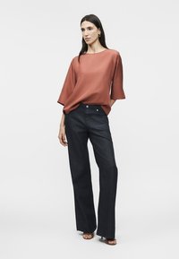 Γυναίκα που φοράει φαρδιά μπλούζα σε σκουρόχρωμο σκουριά με μανίκια τριών τετάρτων, σκούρα τζιν με φαρδιά πόδια και ανοιχτά ψηλοτάκουνα, στέκεται μπροστά σε απλό φόντο.