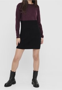 Strickkleid mit der oberen Hälfte in Burgunderrot und der unteren Hälfte in Schwarz, langen Ärmeln und einem runden Ausschnitt. Genossen mit schwarzen Stiefeletten.