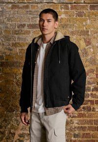 Chaqueta negra con cremallera y capucha forrada de piel sintética beige, llevada sobre una camisa de cuadros y una camiseta blanca, combinada con pantalones cortos de carga ligeros.