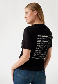 T-shirt in cotone nero con una stampa sul retro che presenta vari nomi di città e date in testo bianco e crema, maniche corte e vestibilità rilassata.