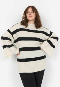 Cremfarbener und schwarzer gestreifter Pullover mit lockerer Passform, weiten Ärmeln und gerippter Textur. Runder Ausschnitt. Kombiniert mit schwarzen Hosen.