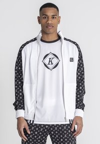 Chaqueta blanca con cremallera y mangas de patrón negro, combinada con una camiseta blanca que lleva un logo negro y pantalones negros con patrón.