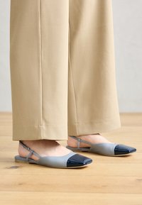 Ballerines grises en cuir avec une cap-toe bleu marine brillant, présentant un design minimaliste et une semelle plate, assorties à un pantalon beige à jambes larges.