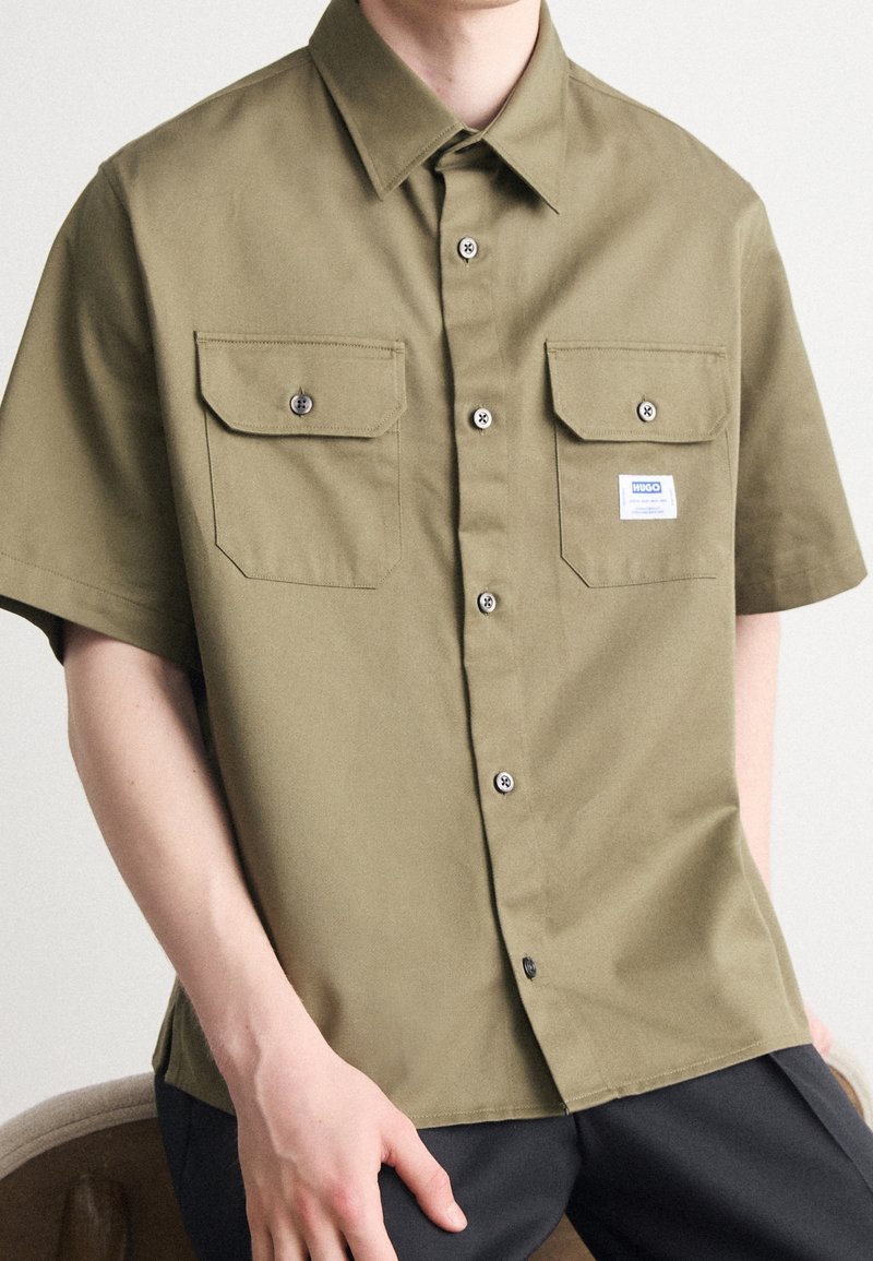Kortærmet skjorte i olivengrøn bomuld med to brystlommer, knapper og et lille mærke. Glat tekstur og afslappet pasform.