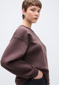 Sötétbarna pulóver bordázott kerek nyakkivágással, nagy méretű fazonban, dombornyomott fekvő felirattal az elején. Puha anyag és lezser ujjvonal növeli a kényelmet.