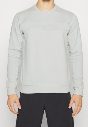 Hombre con sudadera Calvin Klein de manga larga color gris claro y pantalones negros, de pie frente a un fondo blanco liso.