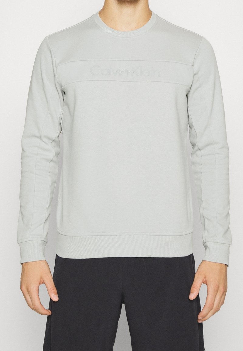 Homme portant un sweat-shirt Calvin Klein à manches longues gris clair et un pantalon noir, debout devant un fond blanc uni.