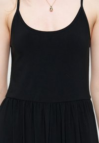 Γυναίκα που φοράει μαύρο αμάνικο φόρεμα με στρογγυλή λαιμόκοψη και χρυσό κολιέ με σταγόνα, δείχνοντας τον πάνω κορμό.