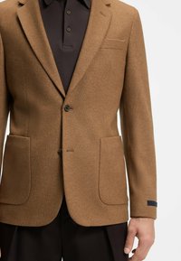 Blazer in lana marrone con due tasche frontali, chiusura a un bottone e rever a punta. Abbinato a una polo scura sottostante.