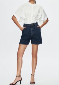 Bílá košile s krátkými rukávy a vysokým pasem modré džínové kraťasy. Modelka má na sobě černé sandály na podpatku.