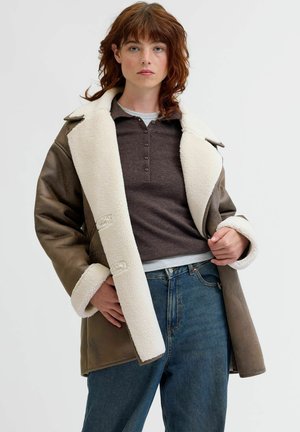 Manteau en shearling marron avec un col en fausse fourrure blanche, doté d'une fermeture à boutons, de manches larges et d'une coupe décontractée, associé à un jean en denim.