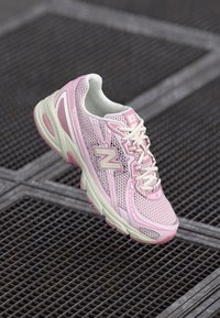 Roosa ja valge New Balance'i jooksujalats, mis hõljub metallvõre taustal, näidates külg- ja ülemisi detaile.
