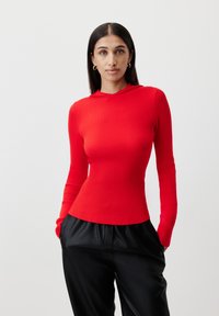 Red ribbed long-sleeve top with a hood; fitted design, soft texture, and a slight flare at the hem. Worn with black loose-fitting pants.