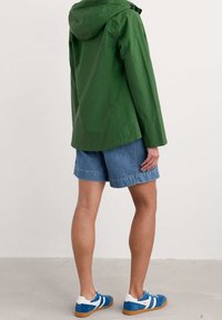 Zelená nepromokavá bunda s kapucí, s hladkou texturou a jednoduchým designem, nošená přes džíny šortky a modré tenisky.
