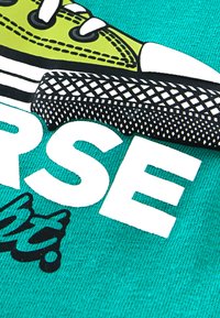 Γραφιστικός σχεδιασμός με πράσινο αθλητικό παπούτσι με λευκά κορδόνια, μαύρη σόλα και σχέδιο από δίχτυ, δίπλα σε έντονο λευκό κείμενο σε τυρκουάζ φόντο.