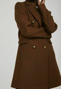 Cappotto marrone in misto lana con design doppiopetto, grandi bottoni in ottone, rever a incavo e tasche laterali. Tessuto lavorato.