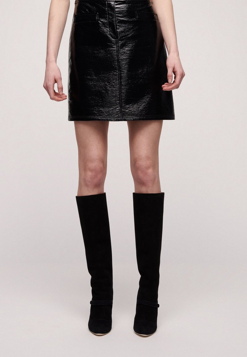 Femme portant une mini-jupe en cuir noire brillante et des bottes noires en daim jusqu'aux genoux, debout les mains détendues le long du corps.