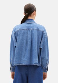Light blue denim jacket with a relaxed fit, buttoned cuffs, and back pleat detail, displayed from the rear, showing a soft texture.