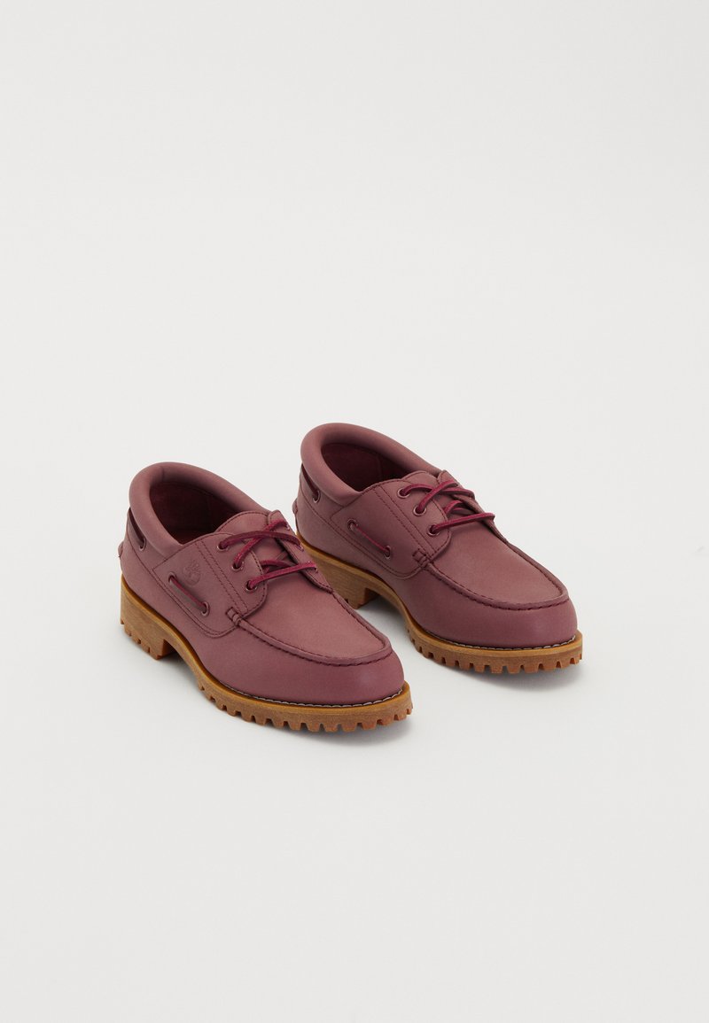 Een paar wijnrode leren bootschoenen met dikke tan rubberen zolen en rode veters op een witte achtergrond.