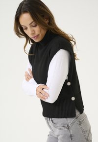 Čierny bezrukávový rolák s rebrovaným lemom, s gombíkovými akcentmi po stranách. Spárovaný s bielou košeľou s dlhými rukávmi pod ním.