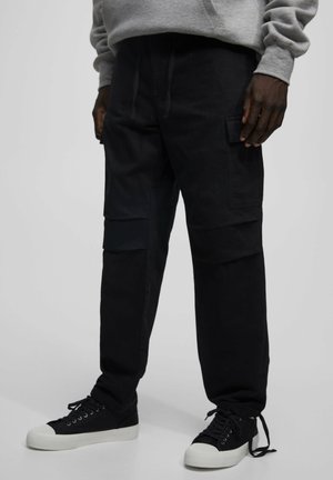 Zwarte cargobroek van stevig katoen met een ontspannen pasvorm, voorzien van meerdere zakken en een trekkoord in de taille, gecombineerd met zwarte sneakers.