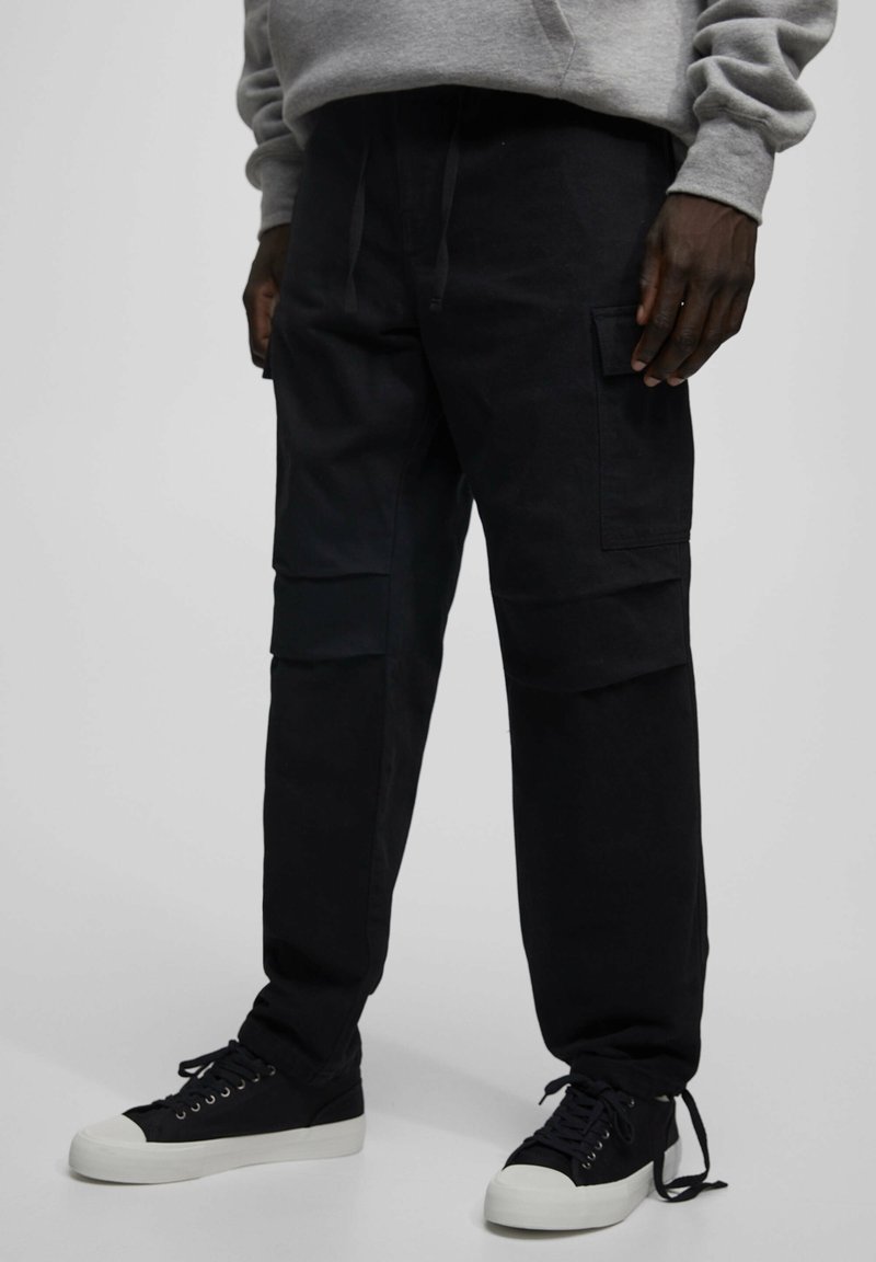 Czarne spodnie cargo wykonane z wytrzymałej bawełny o luźnym kroju, z wieloma kieszeniami i sznurkiem w talii, w połączeniu z czarnymi sneakersami.