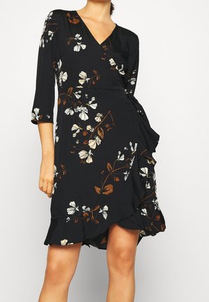 Frau trägt ein schwarzes Wickelkleid mit weißem und braunem Blumenmuster, dreiviertel Ärmeln und einem Rüschensaum vor einfachem Hintergrund.