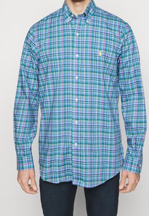 Mann trägt ein langärmeliges Hemd mit grünem, blauem und violettem Karomuster, kombiniert mit dunkler Hose.