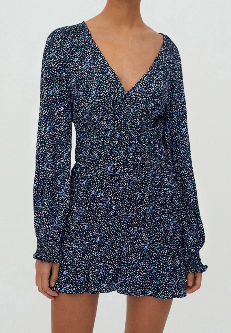 Femme portant une robe courte à manches longues et col en V, avec un fond sombre et un motif floral multicolore de petite taille.