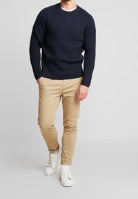 Maglione navy a coste con collo a giro, abbinato a pantaloni slim-fit beige chiaro e sneakers bianche. Il modello è in piedi su uno sfondo neutro.