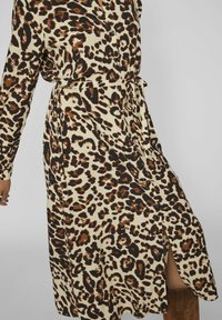 Vestido de estampado de leopardo en beige con patrones marrones y negros, mangas largas, cierre frontal de botones, cintura con cinturón y una abertura alta en el muslo del lado izquierdo.