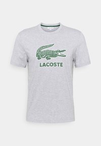 Camiseta de algodón gris con un gran logo de cocodrilo verde y el texto "LACOSTE" debajo. Corte regular con mangas cortas y cuello redondo.
